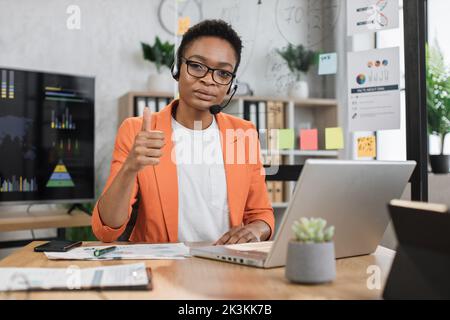 Portrait of attractive african business woman in headset sitting at office desk and looking at camera. Business lady having video conference showing thumb up. Business meeting online. Stock Photo
