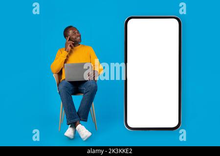 Cheerful black man using laptop, looking at smartphone, mockup Stock Photo