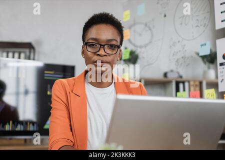Portrait of african business woman in orange suit sitting at desk and looking at camera. Dark haired lady working at modern office with big monitor on background. Stock Photo