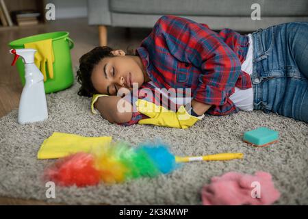 Tired funny young black woman with cleaning supplies sleeping on floor in living room interior Stock Photo