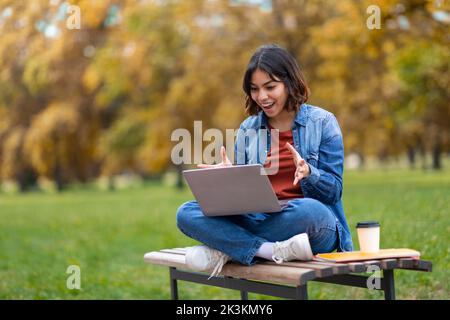 Excited Young Arab Female Student Using Laptop While Sitting On Bench Outdoors Stock Photo