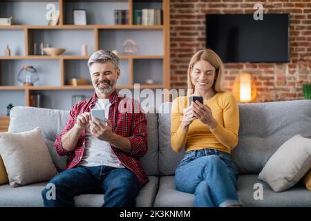 Happy mature european family chatting on smartphones, sit on sofa in cozy living room interior Stock Photo