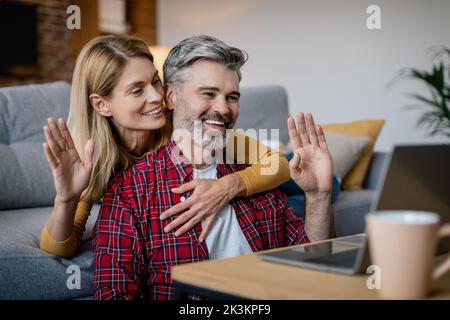 Cheerful mature european lady hugging male, looking at pc, waving hand in cozy living room interior Stock Photo