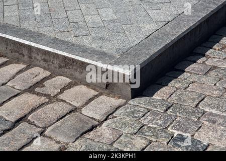 corner of a pedestrian sidewalk made of stone rectangular brick tiles and granite curb dirty in mud near a road paved with rough cobblestone close-up Stock Photo