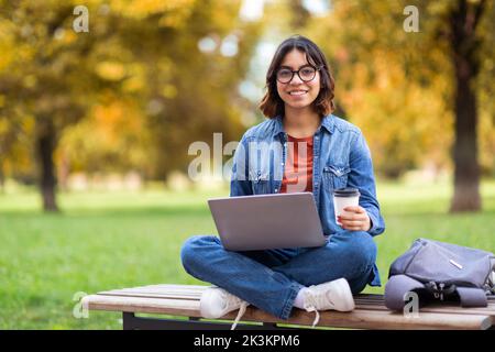 Smiling Young Middle Eastern Female Using Laptop And Drinking Coffee Outdoors Stock Photo