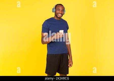 African Man Listening Music Holding Cellphone Wearing Earphones, Yellow Background Stock Photo