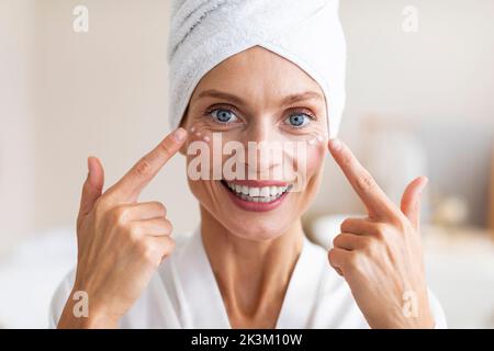 Mature beauty concept. Charming middle aged woman applying face cream under her eyes and smiling at camera Stock Photo