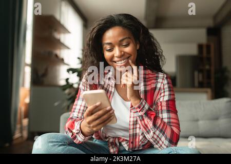 Smiling pretty young african american lady typing on smartphone, chatting in social media alone, sit on sofa Stock Photo