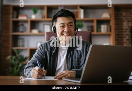 Cheerful middle aged chinese man in headphones has video call on pc, makes notes in cabinet interior Stock Photo