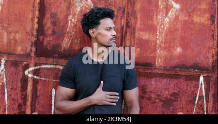 Serious African man standing outside surveilling neighborhood street looking sideways in urban environment Stock Photo