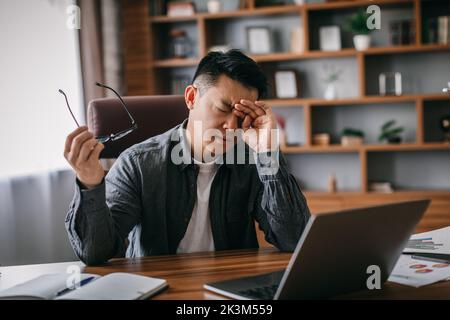 Sad tired adult chinese man takes off his glasses, rubs eyes and rests from work in home office interior Stock Photo