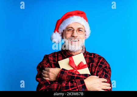 an elderly and well-groomed man in a plaid shirt and santa hat in blue studio background Stock Photo