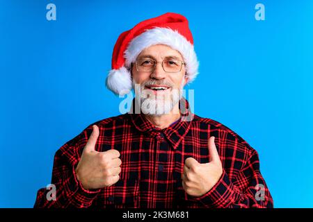 an elderly and well-groomed man in a plaid shirt and santa hat in blue studio background Stock Photo