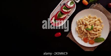 Italian pasta fusilli and caprese salad with tomato, mozzarella and basil with pesto on black rustic background. Extra wide banner. Copy space. Stock Photo