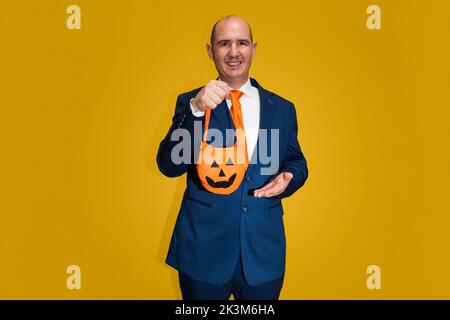 A bald Caucasian adult male dressed in a blue suit, white shirt and orange tie is holding a cloth bag in the shape of a Halloween pumpkin. The backgro Stock Photo