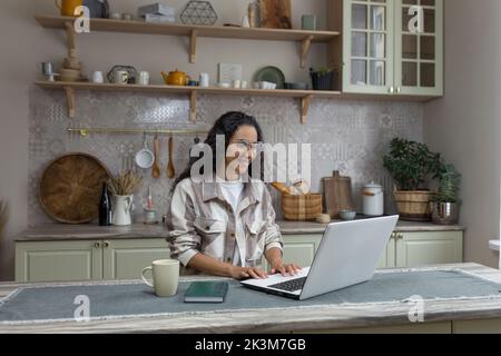 Young beautiful hispanic woman with curly hair working at home using laptop, woman in kitchen smiling and happy remote work freelancer. Stock Photo