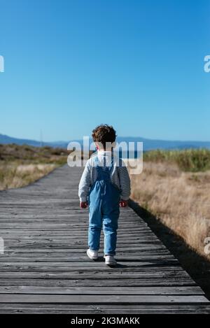 Back view of anonymous preschool kid with curly hair in denim overall walking alone on long paved boardwalk in countryside with clear blue sky Stock Photo
