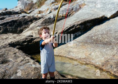 Adorable child fisher in gumboots and overall holding small rod and fishing in shallow clear brook on rocks Stock Photo