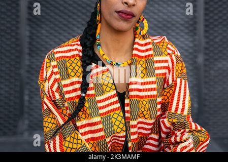 Cropped unrecognizable ethnic woman in colorful shirt with braid near gray wall in daytime on city street Stock Photo