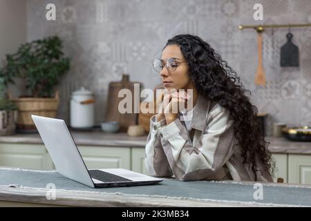 Hispanic woman thinking at home sitting in kitchen working on laptop, freelancer in glasses and curly hair, businesswoman working and learning online remotely. Stock Photo
