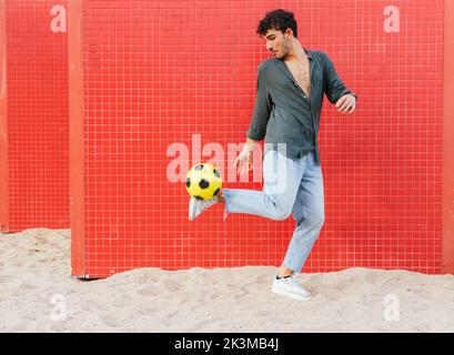 Young Hispanic man in black shirt and jeans kicking and juggling yellow football ball against tiled red wall on street Stock Photo