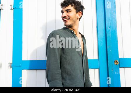 Young smiling Hispanic man in black shirt with curly hair looking away while standing near lumber door Stock Photo