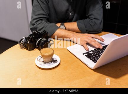 Unrecognizable man using laptop near photo camera and cup of coffee while working on remote project in cafe Stock Photo