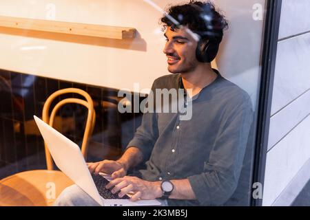 Young smiling Hispanic man in black shirt listening to music in wireless headphones and browsing data on netbook while sitting behind cafe window and Stock Photo