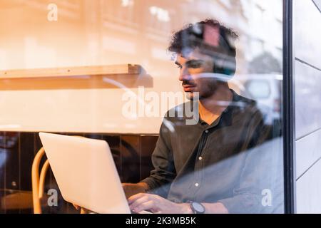 Young Hispanic man in black shirt listening to music in wireless headphones and browsing data on netbook while sitting behind cafe window and working Stock Photo
