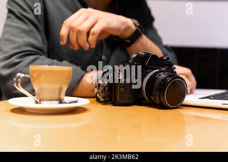 Unrecognizable man using laptop near photo camera and cup of coffee while working on remote project in cafe Stock Photo