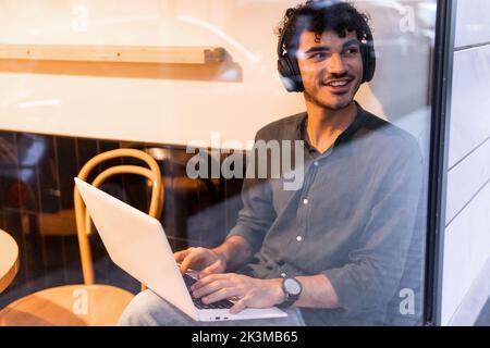 Young smiling Hispanic man in black shirt listening to music in wireless headphones and browsing data on netbook while sitting behind cafe window and Stock Photo