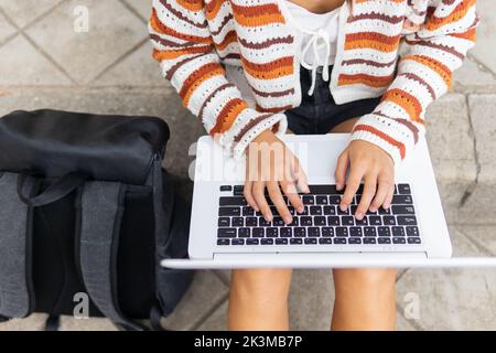 High angle of crop female student browsing netbook and doing homework while sitting on concrete steps near backpack in city Stock Photo