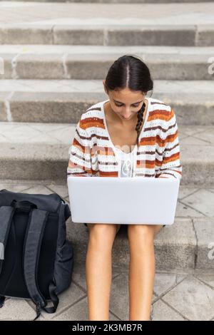 High angle of young female student browsing netbook and doing homework while sitting on concrete steps near backpack in city Stock Photo