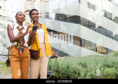 Stylish adult African American traveling female friends with short dark hair using GPS navigator on smartphone and looking away during vacation in mod Stock Photo