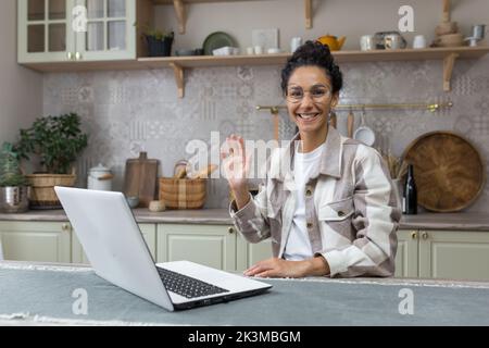 Portrait of young hispanic woman at home, businesswoman smiling and looking at camera waving greeting gesture, freelancer in kitchen using laptop for remote work. Stock Photo