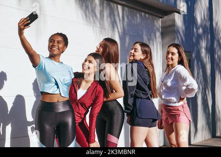 Cheerful young multiracial female best friends in stylish activewear smiling brightly while taking selfie on smartphone after outdoor training Stock Photo