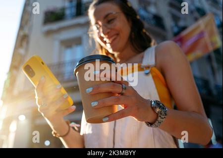 From below soft focus of happy female with paper cup of coffee to go smiling and browsing cellphone on sunny morning on city street Stock Photo