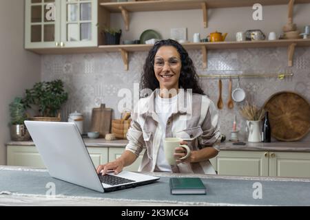 Portrait of young beautiful hispanic woman at home, female freelancer working remotely using laptop, looking at camera and smiling in glasses and curly hair in kitchen. Stock Photo