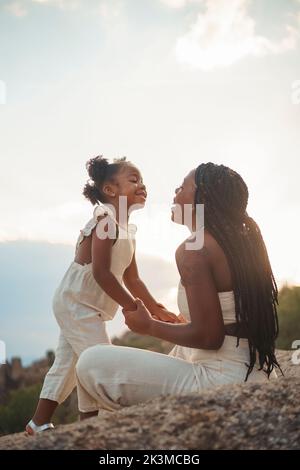 Happy African American mom with curly hair holding hands of daughter and talking while standing on rock against cloudy sundown sky in weekend in natur Stock Photo