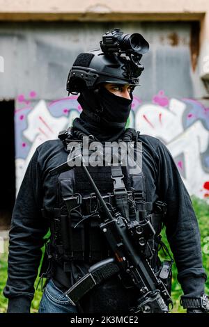 Serious young unrecognizable male warrior in modern protective wear balaclava and helmet with professional rifle looking away while standing near old Stock Photo