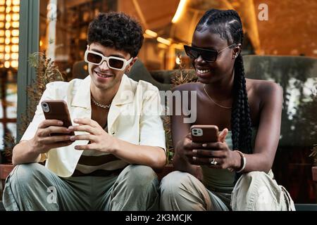 Happy black woman in stylish clothes and sunglasses sitting near boyfriend while using smartphone against glass wall on city street Stock Photo