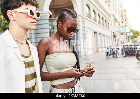 Stylish African American female in sunglasses browsing social media on cellphone while standing near boyfriend on sunny day on city street Stock Photo