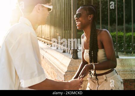 Cheerful black woman in stylish clothes laughing while dancing with boyfriend near road on sunlit city street Stock Photo