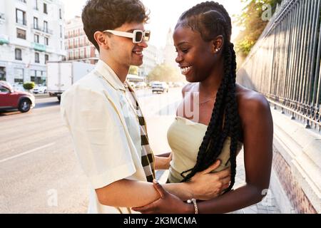 Stylish man embracing cheerful African American woman in while spending time on city street during date Stock Photo