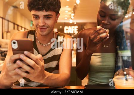 Glad young man using smartphone near black woman biting roll during romantic date in restaurant Stock Photo