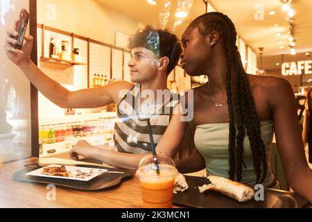 Diverse boyfriend and girlfriend taking selfie via smartphone while having lunch in restaurant together Stock Photo