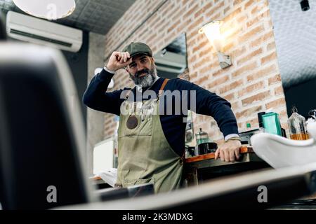 Low angle of cheerful mature male barber wearing cap and apron standing near table in barbershop in loft style and looking at camera Stock Photo
