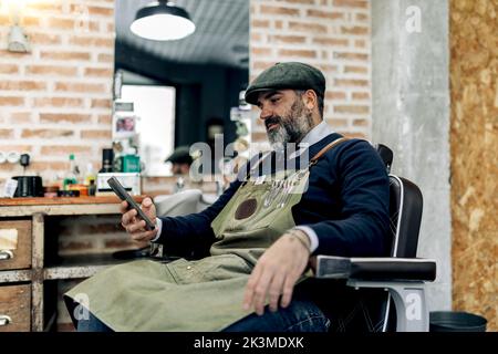 Side view of middle aged male barber sitting on chair in barbershop and browsing mobile phone while having break during work Stock Photo