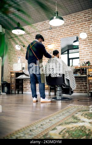 Back view of male customer in cape sitting on chair and getting haircut from professional barber working in grooming salon Stock Photo