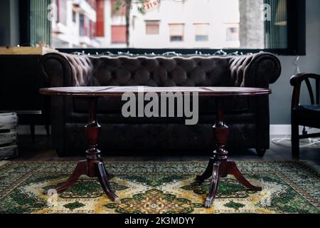 Comfortable vintage brown leather sofa and wooden oval table placed near window in retro styled room with ornamental carpet in daylight Stock Photo
